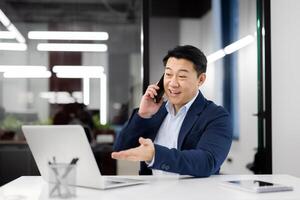 Millennial male sitting alone at workspace and talking by cellphone while spreading hand to portable computer. Friendly manager having remote conversation with colleague about project development. photo
