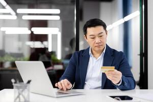 Focused Asian businessman in a modern office making a payment online using a credit card. This professional scene emphasizes e-commerce and secure transactions in a corporate setting. photo