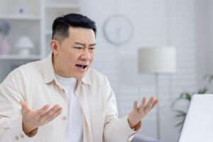 Close-up photo of a young bearded Asian man sitting in the office at a laptop, talking emotionally and unhappily on a call, looking at the screen and gesturing with his hands.