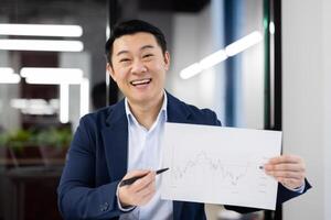 Cheerful taiwan investor showing sheet of paper with growing statistics and pointing with pen to necessary spot. Financial expert felling proud about result of work and made decisions in trading. photo