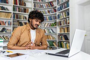 Unhealthy diverse guy looking down and keeping arms on belly while spending day at public library with pc. University student struggling with digestion ache and inability of concentrating on studying. photo