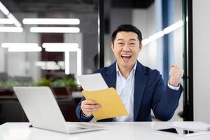 An Asian businessman in a blue suit sits at his desk in a modern office, ecstatically celebrating as he holds an envelope and reads its contents. photo