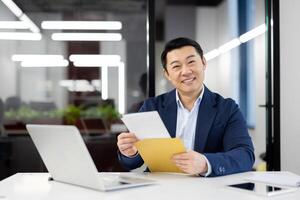 Excited japanese business person smiling happily to camera after reading pleasant information in document. Satisfied male working by desktop with computer and having approval of credit request. photo