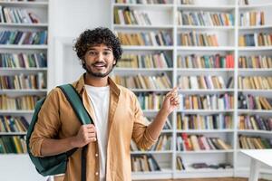 A happy young man with curly hair and a casual outfit is standing in a library. He is carrying a backpack on one shoulder and pointing at something out of frame with a smile on his face. photo