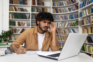 A focused young man is immersed in his studies at the library, surrounded by books, with a laptop for assistance. He seems to be preparing for exams, reflecting the life of a university or academy student. photo