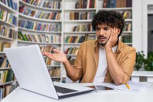 Handsome latino male placing in front of open portable laptop and engaging in online conversation or presentation. confused man gesturing with right hand and discussing something. Home office. photo