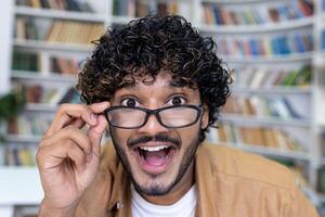 Surprised man looking at camera, hispanic close-up smiling and taking off glasses, student inside academic university library. photo