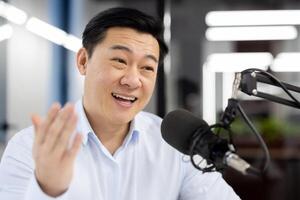 Close-up photo of a young Asian businessman in a white shirt standing in an office and smiling and talking into a microphone while gesturing ,with his hands.