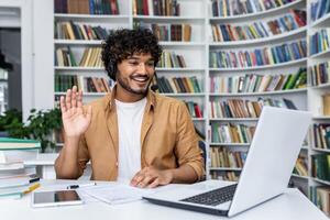 A young man with curly hair and a headset smiles and waves at his laptop camera, engaged in an online call while seated at a desk in a modern library setting with books in the background. photo