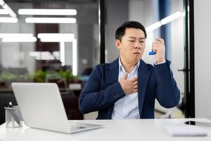 Sick japanese ceo grabbing chest with hand and holding medicine inhaler while sitting by table with portable computer. Asthmatic office manager feeling hard to breath and using emergency drug. photo