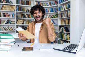 Portrait of young successful student, man received letter of notification of good results of interview exam, hispanic man joyfully looking at camera inside library, holding mail envelope. photo