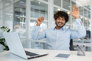Curly islamic man in blue shirt raising arms for stretching back while sitting at workspace with closed eyes. Relaxed employee using free time for exercising and resting from sedentary work. photo