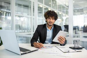 Confident young businessman using a tablet and working on documents in a modern office setting with laptop and papers photo