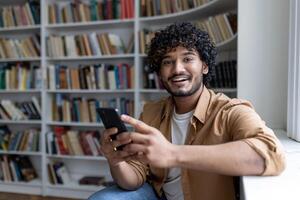 Portrait of young hispanic student, man holding phone smiling and looking at camera, studying inside public library, on university campus. photo
