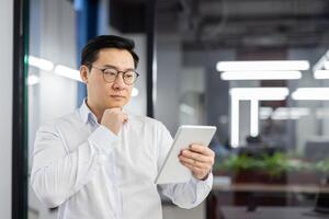 Serious thinking man with tablet computer in hands, asian man at workplace inside office, businessman in shirt reading and analyzing online data using app. photo