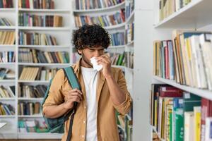 Unhealthy man in casualwear using paper napkin while sneezing and standing by aisle with collection of novels. Sick islamic male struggling with dust allergy in book shop while holding shoulder bag. photo