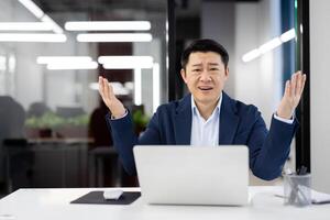 Surprised korean man spreading arms while sitting in front of personal laptop at desk in well-lit office. Evil male solving problems with help of modern technology indoors. photo