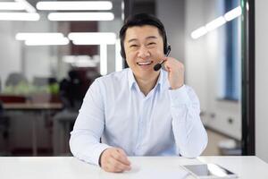 Webcam view, Asian support worker in shirt with headset smiling and looking at camera, businessman working inside office, consulting clients remotely, remote meeting with colleagues partners. photo