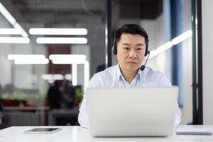 A professional man in a headset is focused on his laptop screen while working in a modern office environment with soft lighting. photo