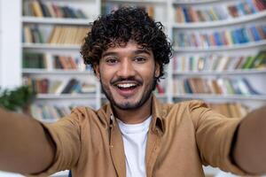 Young Hindu student programmer inside academic library taking selfie photo and talking on call, man with curly hair smiling and looking at camera.