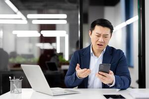 Sad disappointed asian man at workplace, dissatisfied employee in business suit with phone in hands, businessman received online notification message with bad news on smartphone, boss confused. photo