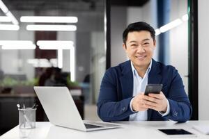 Optimistic asian man holding mobile phone while looking at camera by desktop with pc in modern office interior. Self-employed person having break from work while checking messengers and social media. photo