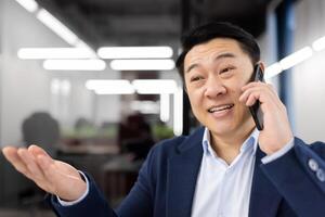 alegre asiático empresario comprometido en un teléfono conversacion en un moderno oficina ajuste. él aparece contento y entusiasta, gesticulando con su mano. foto