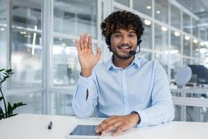 Hindu male wearing headset with microphone and raising hand in greeting gesture while sitting by desk with tablet. Optimistic support operator welcoming new colleague in sharing working space. photo