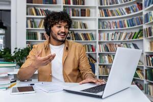 Optimistic indian student using wireless headset and laptop while sitting by desk in library. Confident curly guy answering teacher question while gesturing with hand and looking at computer screen. photo