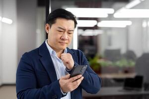 Man in business suit inside office with phone in hands, mature businessman received online notification message with bad news, boss uses app on smartphone, upset received rejection error. photo