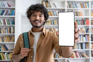 Young successful student studying inside university academic library man smiling and looking at camera showing smartphone screen with white background writing space hispanic man with curly hair. photo