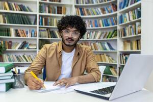 Portrait of a young Indian male student writing an outline while sitting in a university library at a table with a laptop. photo