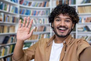 Student call, hispanic man smiling and looking into phone camera, man using app for online communication with friends, student taking selfie photo in library.