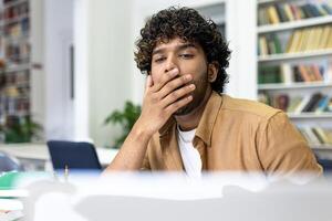 A pensive young man with curly hair concentrates on his studies amidst a library setting, preparing for university exams, reflecting a common academic experience. photo