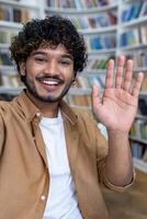 A young happy Spanish male student is talking on a call from a phone in the library. Greets and waves at the camera, participates in the international student exchange program. Vertical photo. photo