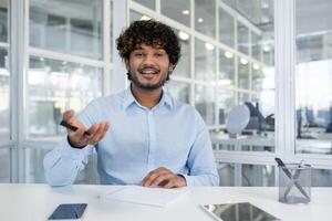 Indian male spreading hands with pen to camera while sitting in front of table with notebook. Smiling hr manager asking question from candidates for hiring and making remarks in copybook. photo