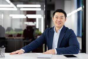 Confident Asian businessman smiling at the camera while sitting at a desk in a contemporary office setting with tech devices. photo