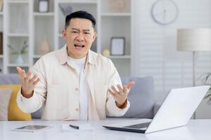 Man expressing frustration during a call meeting at home. He is seated at a desk with a laptop and paperwork. photo