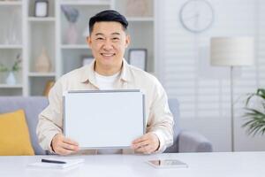 Portrait of a smiling Asian man sitting at a desk in a modern light office and holding a white blank clipboard in his hands, pointing and looking at the camera. photo