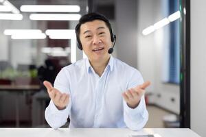 Webcam view, Asian support worker in shirt with headset smiling and looking at camera, businessman working inside office, consulting clients remotely, remote meeting with colleagues partners. photo