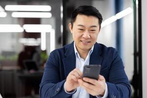 Asian man inside office with phone in hands, smiling contentedly, businessman using smartphone sitting at desk, mature boss in business suit browsing social media, typing text message call. photo