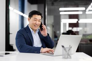asiático empresario en un contento conversacion terminado un teléfono llamar, ordenador portátil abierto, a un bien iluminado moderno oficina espacio. foto