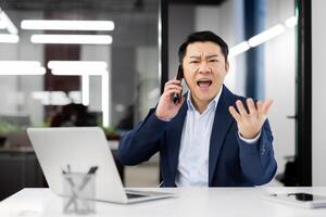 Angry chinese man screaming while having conversation on wireless smartphone on background of glassy office wall. Evil businessman using modern gadgets to staying in touch with colleagues. photo