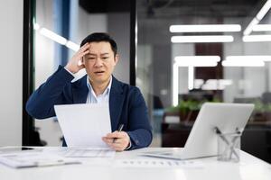 Frustrated korean boss of company grabbing head with hand while reading document in personal cabinet with laptop. Desperate chef receiving unexpected resignation letter from indispensable specialist. photo