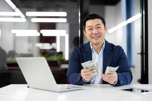 A cheerful Asian businessman counting US dollars at his desk in a well-lit modern office. He exudes success and financial security with a joyful expression. photo