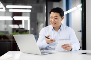 Smiling japanese male entepreneur having online call on modern laptop while holding notebook and pen in hands. Positive man making notes during conversation with interlocutors at office. photo
