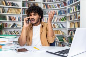 Man in casual work attire gesticulating in anger while talking on the phone, sitting at a desk with a laptop and books. photo