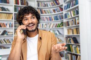 A man in a casual outfit is engaged in a phone conversation, surrounded by bookshelves filled with various books in a well-lit library setting. The concept captures communication and learning photo