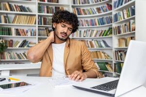 Suffering middle eastern male touching neck with hand while sitting by desktop with gadgets on background of bookshelves. Exhausted man struggling with results of seat with incorrect posture. photo