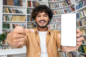 Man smiles brightly as he confidently presents a blank smartphone screen. Perfect for mockups, the background is filled with a variety of books, implying a setting of knowledge and research. photo